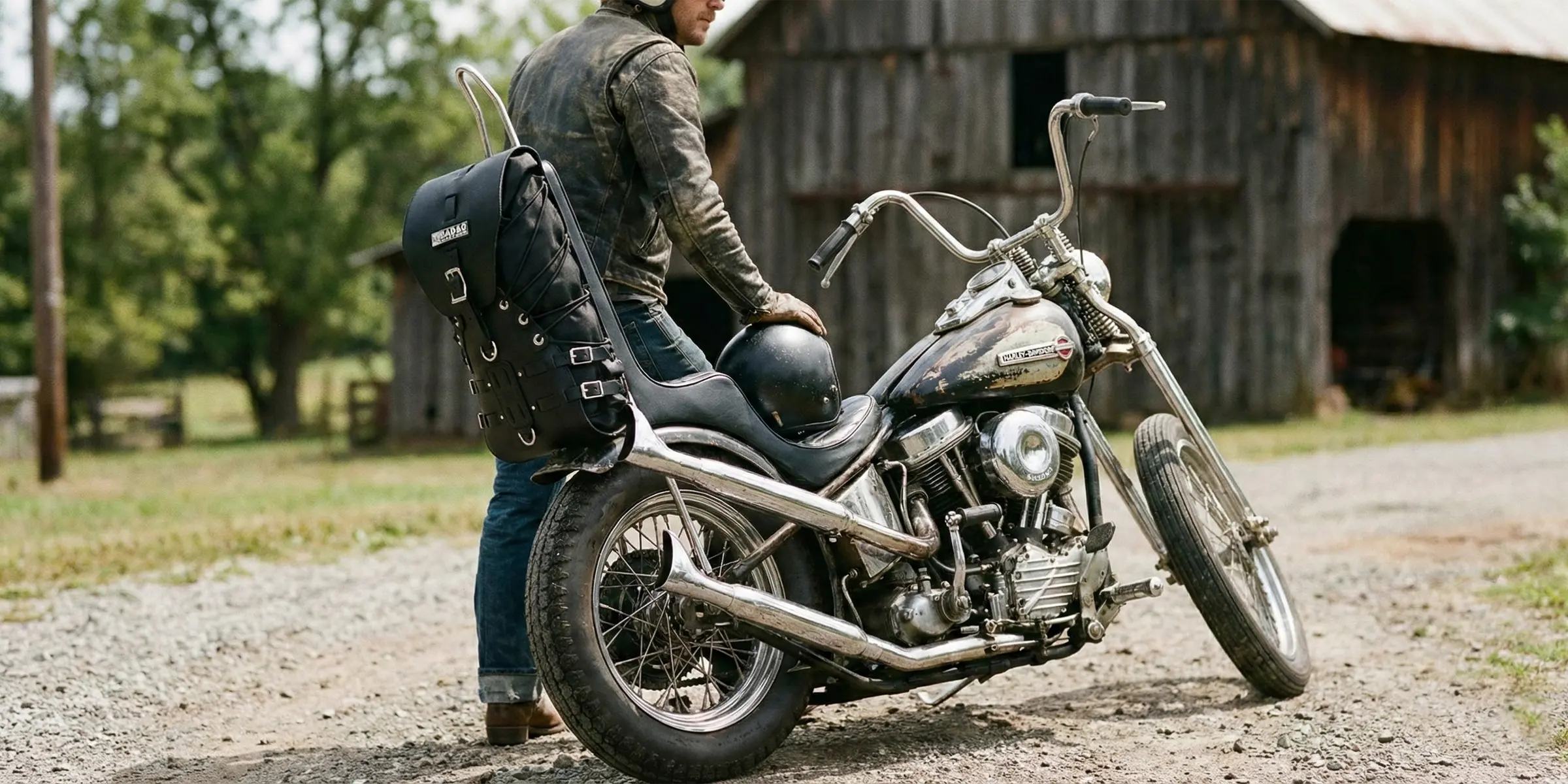 Person sitting standing next to a motorcycle with a barn in the background